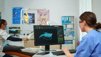 Nurse working at digital dental fingerprint of patient, while specialist dentist doctor with face mask is speaking to man with toothache sitting on stomatological chair preparing tools for examination. photo