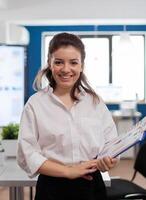 Project manager looking at camera smiling, standing in middle of conference room while partners talking in background. Manager working in professional start up financial business, modern company workplace ready for meeting. photo
