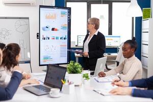 Elderly project manager pointing at desktop presenting statistical data, briefing diverse group of employees. Multiethnical businesspeople working in professional startup financial office during conference meeting photo