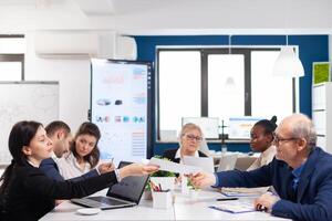 Young employee exchanging documents with senior executive in startup board room with multiethnic colleagues coworkers analysing graphs and charts in briefing corporate. photo