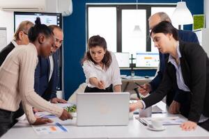 Diverse team of workers talking, looking at documents and analyzing graphs data standing at conference desk. Multiethnic colleagues working planning success financial strategy discussing in office photo