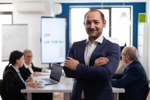 Young man entrepreneur smiling in front of camera standing in brainstorming room, preparing for meeting with partners. Manager working in professional start up financial business, modern company workplace ready for meeting. photo