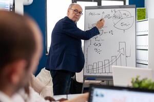 Elderly businessman giving presentation to multiethnic team of colleagues using flip chart. Serious speaker boss executive, business trainer explaining development strategy to motivated mixed race employees. photo