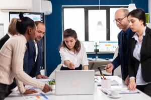 Diverse team of workers talking, looking at documents and analyzing graphs data standing at conference desk. Multiethnic colleagues working planning success financial strategy discussing in office photo