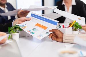 Close up view desk full of papers stats shown on charts and diverse businesspeople. Colleagues working in office at conference table in startup management company. photo