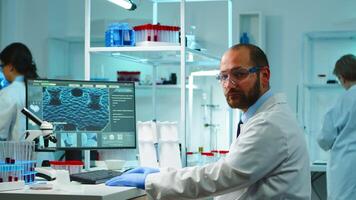 Worn out doctor typing on computer and looking exhausted at camera in equipped lab late at night. Medical chemists examining virus evolution using high tech for scientific research, vaccine development. photo