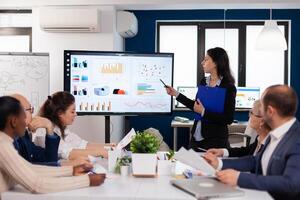 Young team leader in big corporation briefing coworkers pointing at graph meeting. Corporate staff discussing new business application with colleagues looking at screen photo
