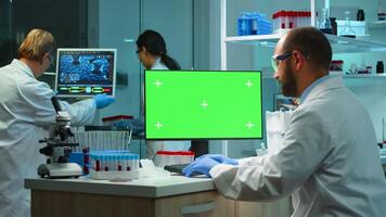 Man researcher looking at chroma key computer in modern equipped lab working overtime. Team of microbiologists doing vaccine research writing on device with green screen, isolated, mockup display. photo