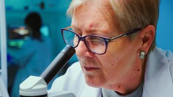 Close up of senior scientist making research using microscope while colleagues working on background in modern equipped laboratory. Stuff analysing vaccine evolution using high tech photo