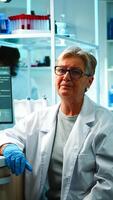 Portrait of old chemist woman with fatigued face in front of camera in equipped lab late at night. Team examining virus evolution using high tech and chemistry tools for research, vaccine development photo