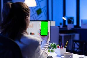 Entrepreneur looking at smartphone with green screen sitting at office desk overworking. Manager project watching desktop monitor display with mockup chroma key during videocall meeting remote photo