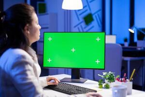 Businesswoman looking at green screen monitor of computer sitting at desk during online web conference. Freelancer watching desktop monitor display with green mockup, chroma key in business office photo