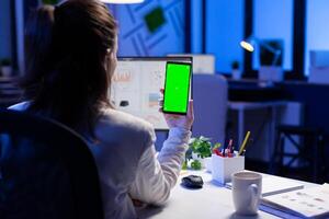 Freelancer looking at mobile phone with green screen display during online videocall meeting sitting at desk in business office. Businesswoman watching desktop monitor with green mockup, chroma key photo