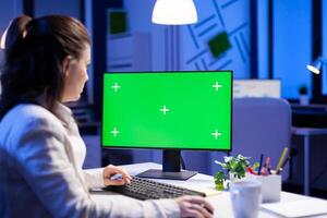 Entrepreneur working in front of green screen display sitting at desk during videocall meeting in business office. Businesswoman watching desktop monitor with green mockup, chroma key, working overtime. photo