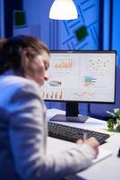 Tired entrepreneur trying to finish business project during internet videocall in front of computer, taking notes on notebook. Woman employee sitting at desk in workplace working at marketing ideas photo