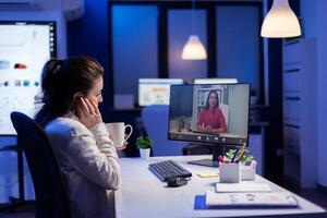 Businesswoman discussing with corporate partner sitting in front of computer in start-up office during virtual web meeting late at night, using wireless headphone and modern technology network photo