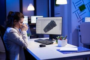 Woman engineer working late at night on 3D model of industrial turbine while drinking coffee in front of computer. Workaholic freelancer studying prototype design on pc showing cad software on device display photo