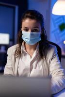 Close up of businesswoman with face mask checking emails late at night in new normal business office before deadline. Taking notes, analysing documents overtime during global pandemic photo