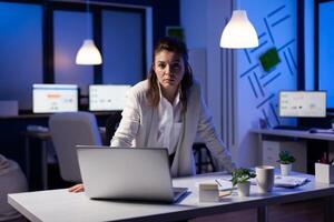Business woman looking tired at camera standing near desk in start-up business company late at night. Focused entrepreneur using technology network wireless for developing new marketing system photo