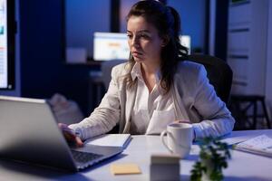 Project manager talking online with coworkers late at night sitting in busineess office on professional laptop. Overworked employee using technology network wireless speaking on virtual meeting photo