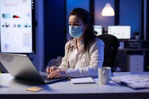 Manager woman with face mask working overtime in new normal business office looking at team project on professional laptop. Analysing financial documents sitting at desk during global pandemic photo