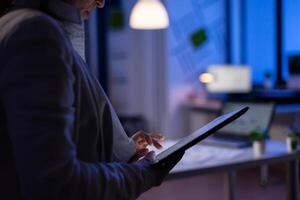 Close-up of woman hands typing on tablet checking financial graphs standing in start-up office late at night. Manager using social network, texting and blogging working overtime for job photo