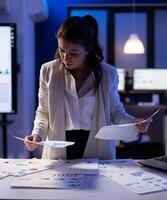 Stressed manager woman working with financial documents checking graphs, holding papers late at night. Business woman standing in start-up office doing overtime work to respect deadline. photo