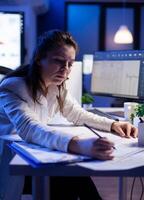 Woman architect analysing and matching blueprints for new building project sitting at desk. Graphic designer using arhitecture prototype plans working overtime in corporate office late at night photo