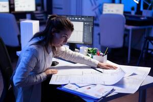 Overworked woman architect checking and matching blueprints sitting at office desk in front of computer. Perfectionist designer using arhitecture blueprints of buildings creating industrial prototype photo