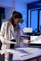 Busy business woman looking on financial reports, checking statistics graphs standing at desk late at night in start-up office. Manager using social network working on marketing project photo