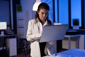 Tired entrepreneur analysing financial reports standing in start up office holding professional laptop. Focused employee calculate marketing profit late at night using modern technology network photo