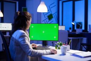 Entrepreneur working in front of green screen display sitting at desk during videocall meeting in business office. Businesswoman watching desktop monitor with green mockup, chroma key, working overtime. photo