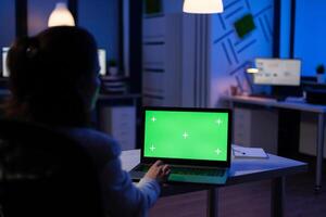 Back view of business woman looking at green screen laptop sitting at desk in start up company office . Exectutive manager watching desktop monitor display with green mockup, chroma key photo