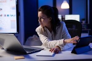 Manager woman using laptop and tablet in same time working on financial reports in business start-up office . Busy multitasking employee using modern technology network wireless late at night photo