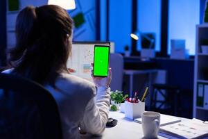 Entrepreneur looking at smartphone with green screen sitting at office desk overworking. Manager project watching desktop monitor display with mockup chroma key during videocall meeting remote photo