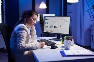 Tired entrepreneur trying to finish business project during internet videocall in front of computer, taking notes on notebook. Woman employee sitting at desk in workplace working at marketing ideas photo
