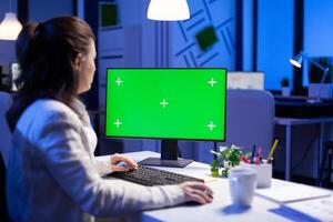 Businesswoman looking at green screen monitor of computer sitting at desk during online web conference. Freelancer watching desktop monitor display with green mockup, chroma key in business office photo