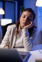 Exhausted businesswoman looking tired in camera sighing resting head in palm late at night in business office. Focused entrepreneur using technology network wireless, working at marketing statistics photo