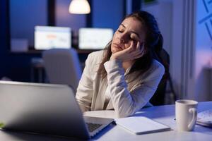 Exhausted freelancer falling asleep in front of laptop checking financial reports in company office late at night. Manager using modern technology network wireless working on economic statistics photo