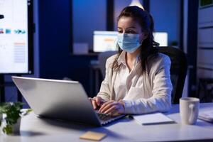 Close up of businesswoman with face mask checking emails late at night in new normal business office before deadline. Taking notes, analysing documents overtime during global pandemic photo
