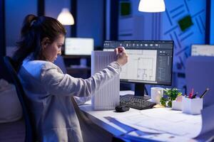 Woman architect analysing and matching blueprints for new building project sitting at desk. Graphic designer using arhitecture prototype plans working overtime in corporate office late at night photo