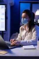 Manager woman with face mask working overtime in new normal business office looking at team project on professional laptop. Analysing financial documents sitting at desk during global pandemic photo