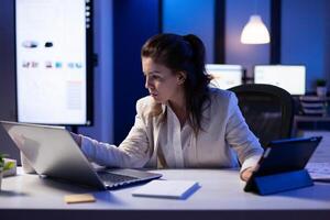 Manager woman using laptop and tablet in same time working on financial reports in business start-up office . Busy multitasking employee using modern technology network wireless late at night photo