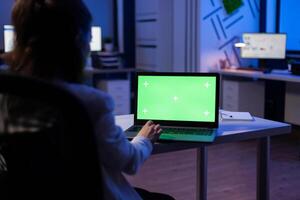 Back view of business woman looking at green screen laptop sitting at desk in start up company office . Exectutive manager watching desktop monitor display with green mockup, chroma key photo