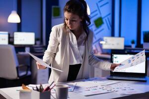 Busy business woman looking on financial reports, checking statistics graphs standing at desk late at night in start-up office. Manager using social network working on marketing project photo