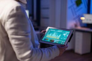 Close-up of woman hands typing on tablet checking financial graphs standing in start-up office late at night. Manager using social network, texting and blogging working overtime for job photo