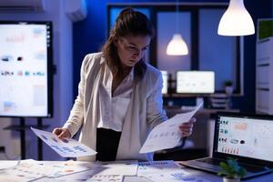 Busy business woman looking on financial reports, checking statistics graphs standing at desk late at night in start-up office. Manager using social network working on marketing project photo