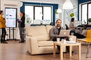 Portrait of man employee typing on laptop looking at camera smiling while diverse team working in background. Multiethnic coworkers talking about startup financial company in modern business office photo