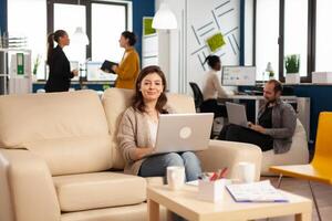 Portrait of entrepreneur typing on laptop looking at camera smiling while diverse team working in background. Multiethnic coworkers talking about startup financial company in modern business office photo