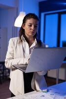 Tired entrepreneur analysing financial reports standing in start up office holding professional laptop. Focused employee calculate marketing profit late at night using modern technology network photo